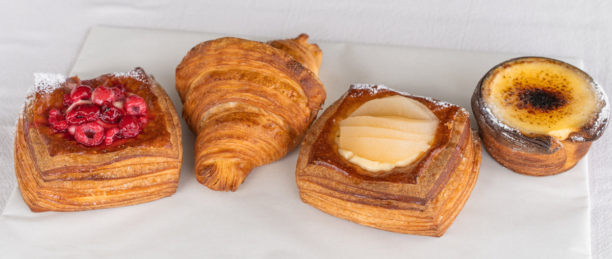 Four assorted pastries arranged on a white plate against a white background: a square pastry topped with red berries, a croissant, a square pastry with a pear filling, and a small burnt muffin or crème pastry.