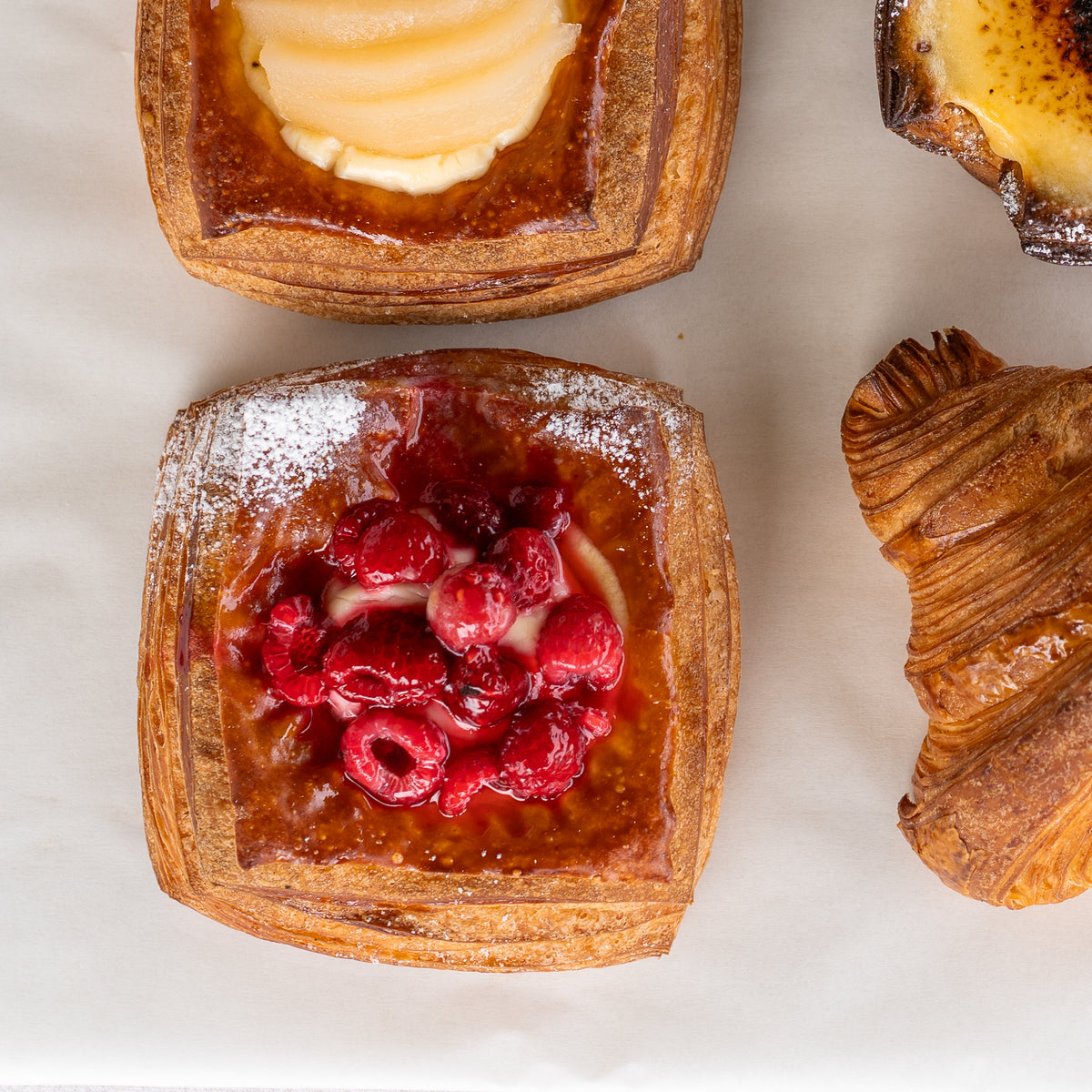 Top-down view of assorted pastries on white parchment paper, including a square flaky pastry topped with red raspberries.