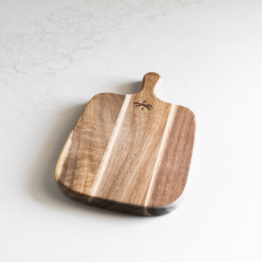A rectangular wooden serving board with rounded corners and a small handle, featuring natural wood grain and a small engraved logo.