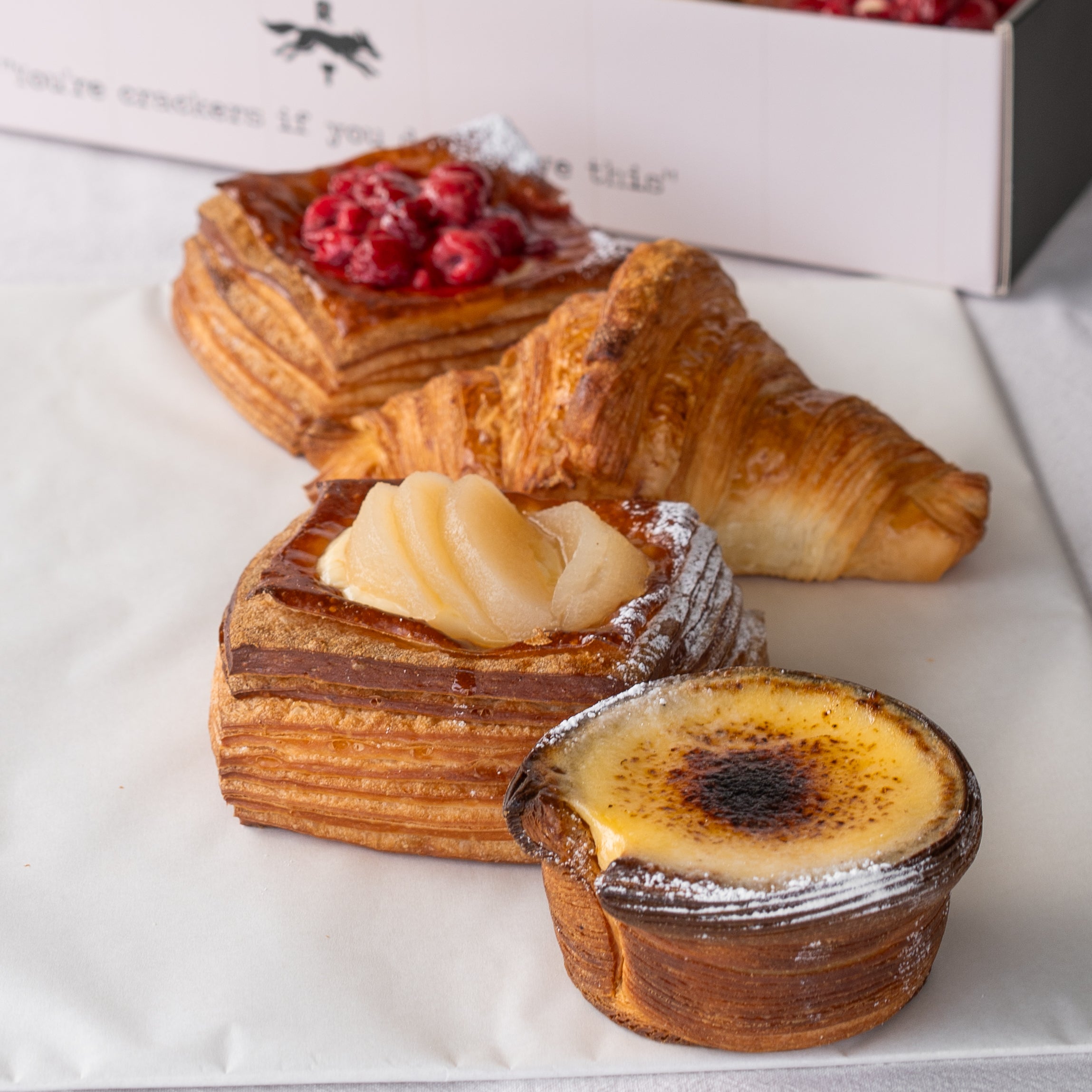 Four assorted baked pastries arranged on white parchment paper, including a croissant, a pastry topped with red berries, a layered pastry with sliced fruit, and a small burnt-mustard tart, with part of a branded box visible in the background.