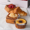 Four assorted baked pastries arranged on white parchment paper, including a croissant, a pastry topped with red berries, a layered pastry with sliced fruit, and a small burnt-mustard tart, with part of a branded box visible in the background.