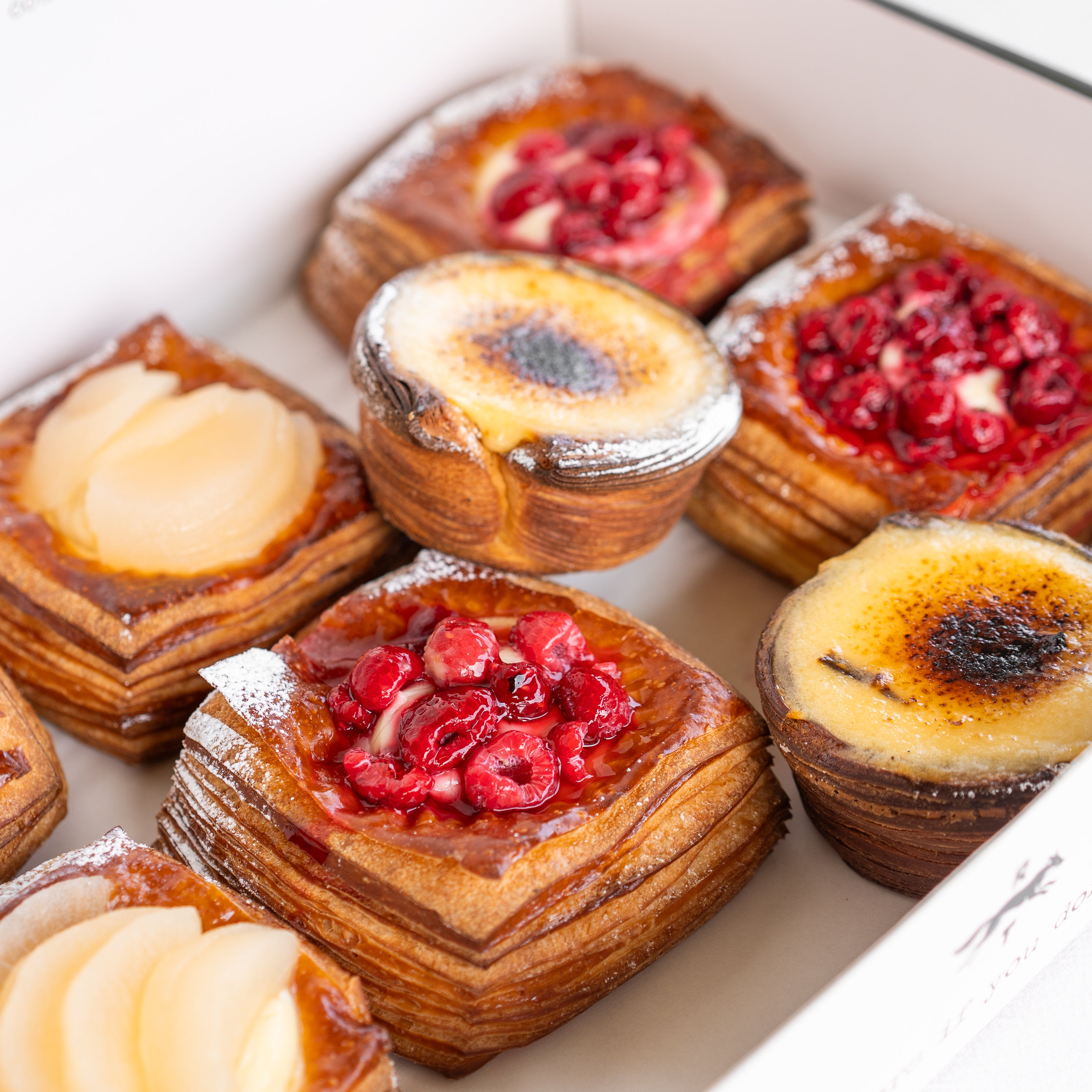 An assortment of baked pastries arranged in a white box, including square flaky pastries with pear slices, round pastries with berry toppings.