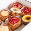 An assortment of baked pastries arranged in a white box, including square flaky pastries with pear slices, round pastries with berry toppings.